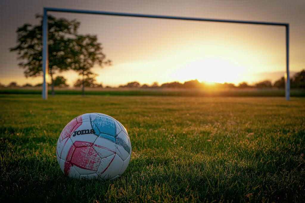 Un balón en el césped de un campo de fútbol al atardecer, con la portería al fondo